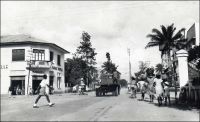 Douala, carrefour avenue Poincaré et rue du 27 août. – « Le voyageur qui débarque pour la première fois à Douala est surpris de se trouver devant un vaste chantier […]. Dans les rues – goudronnées depuis peu – le mouvement des automobiles et bicyclettes devient tel que le service de la circulation a dû installer aux principaux carrefours des « clignotants » vert et rouge, les agents, les « polices » comme on les appelle ici, ne parvenant plus, par leurs seuls moyens individuels, bâton et sifflet, à éviter, aux heures d’affluence, des embouteillages spectaculaires ». Sources : « Douala, porte du Cameroun » (brochure), Douala, Secrétariat social du Cameroun et Chambre de commerce, d’agriculture et d’industrie du Cameroun, 1953.