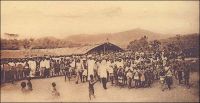 Cameroun français, en brousse, arrivée triomphale du Missionnaire au village. – « [Le Cameroun compte] en tout, une soixantaine de pères et 2500 catéchistes pour 232 000 catholiques, 142 000 catéchumènes ou 374 000 Noirs plus ou moins christianisés, soit en moyenne, un prêtre pour 6500 fidèles ou demi-fidèles et un catéchiste pour 150. […] Dans la brousse, on n’est à son presbytère que la moitié du temps. Ainsi, à Edéa, le supérieur peut en quinze jours visiter vingt-cinq de ses cinquante postes ; au sud et au sud-est, les derniers sont à cinquante ou soixante kilomètres ; faute de bonnes routes, il doit aller entièrement à pied ; il fait chaque jour plus de cinq lieues dans la région du sud où les villages sont rares, deux heures seulement de marche au sud-est où les habitations sont plus rapprochées. […] Suivant l’état des chemins, tantôt dans une vieille petite Ford, tantôt en motocyclette, tantôt à cheval, tantôt à pied, par des pistes souvent barrées de troncs d’arbres, coupées de ruisseaux, même de rivières, où il arrive qu’on trouve un vieux pont de liane qu’il faut réparer avant de s’y hasarder. ». – Carte postale éditée par la « Mission des Prêtres du Sacré-Cœur de St-Quentin ».   Source : J. Wilbois Le Cameroun, éditions Payot, Paris, 1934.
