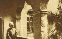 Le sultan de Foumban sur la galerie de son palais. – Dernier représentant de l’empire Bamoun, l’un des plus anciens du continent, le sultan de Foumban embarrasse les  autorités mandataires françaises - cette partie du Cameroun est placée en 1922 sous mandat français par la Société des Nations. Elles vont s’employer à réduire son pouvoir et son emprise, en transformant l’empire en simple région dès 1922, puis l’année suivante, en simple subdivision de la circonscription de Dschang. La même année, le souverain Njoya est déposé. Exilé à Mantoun dans un premier temps, puis à Yaoundé, il meurt en 1933, scellant la disparition de l’organisation politique Bamoun et la suprématie de l’administration coloniale française. – Ce cliché fait partie de l’œuvre du photographe Georges Goethe. Né en 1897 en Sierra Leone, il vient au Cameroun  en 1922 comme agent commercial et, fasciné par les hommes, les villes et les paysages, se lance en amateur dans la photographie. Pionnier du genre, il acquiert vite une réputation, et les chefs de villages le demandent. En 1931, il devient professionnel et ouvre à Douala son studio, appelé « Photo Georges », où seront formés nombre des premiers photographes camerounais. Ses archives constituent un fonds d’une grande richesse sur la vie du pays. Depuis sa disparition  en 1976, son fils, Doualla Cyrille Goethe, poursuit son oeuvre.