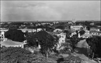 Douala, vue générale. – « A Douala, il y a encore beaucoup d’anciennes « cases » (ici on appelle « cases » toutes les maisons, qu’elles soient occupées par les Européens ou les indigènes) typiquement coloniales ou des cases nouvelles construites sur le même principe ; un rez-de-chaussée surélevé entouré le plus souvent d’une véranda, un toit de tôle ondulée, une cuisine indépendante. Ces cases sont généralement agréables à habiter car elles sont bien aérées. Mais on rencontre aussi des « cases » qui ressemblent aux maisons de France : immeubles à 1 ou 2 étages avec logements ou appartements » (1). Le recensement de novembre 1951 dénombre à Douala une population de 5250 Européens et 115 000 Africains. - On distingue, à l’arrière plan, la cathédrale Saints-Pierre-et-Paul. Source : « Douala, porte du Cameroun » (brochure), Douala, Secrétariat social du Cameroun et Chambre de commerce, d’agriculture et d’industrie du Cameroun, 1951.