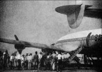 Arrivée de l’avion d’Air France à Douala, en 1950. – « Le camp d’aviation, son bar plutôt, est un des rendez-vous où l’on papote, les arrivées et les départs d’avions fournissant des sujets de conversation inépuisables » (1). Pour se rendre au Cameroun en 1950, les voyageurs doivent posséder un passeport, doivent avoir reçu les vaccinations antiamaryle et antivariolique. De plus, ils doivent verser une caution, ou produire une dispense délivrée par la Sûreté du territoire à la demande de l’employeur. Le montant de la caution s’élève à 30  000 francs CFA pour les Français, Belges, Allemands, Espagnols, Hollandais, Italiens, Luxembourgeois, Portugais et Suisses, à 50 000 f. CFA pour les Russes et Américains du Nord et du Sud, à 10 000 f. CFA pour les « Originaires de la Côte d’Afrique », et 40 000 f. CFA pour les autres nationalités. Les voyageurs doivent aussi disposer d’une autorisation d’embarquement, délivrée par la Délégation du Cameroun en France. Les vols d’Air France entre  Paris et Douala par Constellation durent 14 heures, avec une escale à Alger, et le billet coûte 57 800 f. CFA (115 600 francs métropolitains). Les vols UAT, sur DC4, passent par Alger et Niamey, et durent 18 heures comme les vols TAI qui passent par Alger et Kano, et le billet coûte sur ces compagnies 46 500 f. CFA, soit 93 000 f. métropolitains. Par comparaison, le SMIG (salaire minimal interprofessionnel garanti) créé en France en 1950, ne s’élevait alors qu’à 64 francs métropolitains par mois !  Source : « Douala, porte du Cameroun » (brochure), Douala, Secrétariat social du Cameroun et Chambre de commerce, d’agriculture et d’industrie du Cameroun, 1951.