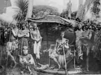 Cameroun français, un Chef en tenue de gala avec son fon masqué. Carte éditée par la mission des prêtres du Sacré-cœur de St-Quentin. - Sur cette photo, probablement prise dans les années 1920-1930, on voit un grand chef (fo) Bamiléké de l'Ouest-Cameroun, revêtu de ses vêtements d'apparat en tissu ndop (batik de coton, teint à l'indigo) et notamment sa grande coiffe de plumes (ten), son baudrier, et son immense pipe 