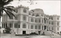 Abidjan, Palais du Gouvernement. – Ce palais rococo se dressait à l’emplacement de l’actuelle Présidence de la Côte d’Ivoire. Symbole du pouvoir colonial, il fut démoli après l’indépendance et remplacé par un bâtiment sans grand caractère. Sa construction avait été envisagée dès 1924, et pour la première fois on avait confié le projet à un cabinet d’architectes, Jaussely et Olivier qui était spécialisé dans l’architecture coloniale. Précédemment, la réalisation de tous les édifices officiels de Côte d’Ivoire était assurée par les Travaux Publics, un service de l’administration coloniale. Ce premier recours à une signature architecturale correspond à un revirement dans la politique antérieure du moins coûtant, tandis que cette colonie ne cesse de prospérer. Le chantier ne fut effectivement entamé qu’en 1931. Il avait finalement était confié à l’architecte Baudouin qui résidait à Abidjan. La construction, qui dura deux ans, ouvrait l’ère des grands travaux préparant le transfert de la capitale à Abidjan – elle se trouvait précédemment à Bingerville - en 1933. La richesse des décorations et installations du palais étaient alors inédite en Côte d’Ivoire.