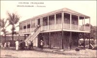 Grand-Bassam, factorerie. – Le commerce de traite, qui prévaut dans le système colonial, consiste à échanger des marchandises manufacturées occidentales contre des matières premières tropicales, lesquelles sont ensuite revendues sur le marché métropolitain. Selon les époques et les lieux, ces échanges utilisent la monnaie, ou s’en affranchissent. La factorerie est la base locale de ce négoce. La première factorerie de Côte d’Ivoire aurait été créée en 1863 par le rochelais Arthur Verdier, à Assinie, où résidait alors le représentant de la France. Le peu d’empressement des grandes compagnies commerciales à développer leur réseau en Côte d’Ivoire, laissa la place à de petits commerçants qui maillèrent progressivement le territoire. A partir de 1858, les négociants étrangers furent d’ailleurs autorisés à s’installer dans les comptoirs français. La factorerie à proprement parler (on ne voit ici que le débarcadère) n’est bâtie en dur que lorsqu’elle s’est avérée viable commercialement, disposant d’un réseau de sous-factoreries dans l’intérieur du pays. La construction comprend alors un solide rez-de-chaussée, pour protéger le stock, aux épais murs de maçonnerie de pierre ou de briques importées, et avec de petites ouvertures à barreaux. L’habitation du traitant est aménagée à l’étage, dans un bâti plus léger, et entourée d’une galerie.