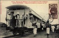 Voyage du ministre des colonies à la Côte d’Afrique. Départ d’Abidjan en chemin de fer. - Il s’agit du déplacement de Raphaël Milliès-Lacroix, ministre des colonies dans le premier gouvernement Clemenceau, qui s’embarqua le 13 avril 1908 à Lisbonne sur un navire des Messageries Maritimes et visita successivement le Sénégal, la Guinée française, la Côte d’Ivoire et le Dahomey. Ce voyage, sous le signe de l’étude plus que du protocole (outre le ministre, la délégation ne comptait que deux collaborateurs), rencontra un chaleureux accueil des colons. Il fut couvert par le photographe Edmond Fortier de Dakar qui rejoignit la mission dès cette première escale.  Les travaux du chemin de fer « Abidjan-Niger » débutent en 1904. En 1909, la ligne rallie Dimbokro qui reste le terminus jusqu’à l’ouverture en 1912 d’un nouveau tronçon de 181 km, joignant Bouaké. Puis, entre 1919 et 1923, les 55 km séparant Bouaké de Katiola sont couverts. Entre 1924 et 1929 le tronçon Katiola-Ferkéssédougou, long de 187 km, est construit ; les 238 km supplémentaires, permettant de rallier Bobo-Dioulasso, sont achevés en 1932. La jonction ferroviaire entre Abidjan et Ouagadougou est réalisée en 1954. Enfin, du temps de la révolution Burkinabè, un réel effort populaire permet la construction d’un tronçon supplémentaire d’une centaine de kilomètres jusqu’à Kaya, à l’instigation de Thomas Sankara lui-même. Ce dernier tronçon n’a jamais été exploité. Ainsi, lorsque fut prise cette photo en 1908, le train n’allait pas au-delà du N’zi, une rivière proche de Dimbokro, qui sera franchie l’année suivante grâce à l’édification d’un imposant viaduc métallique. Et quand Marcel Paternot, l’auteur des lignes qui suivent et décrivent bien le train, l’emprunte peu avant la seconde guerre mondiale, le terminus est Bobo-Dioulasso. - « Il fait à peine jour que la gare [d’Abidjan], une construction bien quelconque qui rappelle le type standard des petites stations de la Métropole, est déjà très animée. Le train est rangé sur le quai et une foule d’indigènes aux pages multicolores s’agitent de toutes parts. Nous prenons place en seconde classe où nous trouvons déjà quelques noirs. Ça ne vaut pas nos voitures d’Europe, mais enfin c’est convenable, genre train départemental, compartiments ouverts sans porte sur le couloir latéral, petites plateformes en avant et en arrière » (1). Source : (1) Source : Paternot, Marcel, Lumière sur la Volta – Chez les Dagari, Lyon, Editions de la plus grande France, 1946.