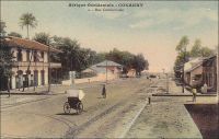 Conakry, Rue Commerciale - Également connue comme rue, avenue ou boulevard du Commerce ou encore « 3ème boulevard », cette artère majeure de la capitale guinéenne aboutit au port. En 1899, l’administrateur Bie la décrit en ces termes : « n’a pas moins de 30 m. de large sur une longueur d’environ un kilomètre. De larges trottoirs bordant cette rue, de splendides maison à étage, toutes neuves, lui donnent une apparence toute moderne » (1). Le chemin de fer Decauville, que l’on distingue sur l’image, fut déployé dans ce quartier de négoce à partir de 1897. Cette installation, constituée de rails écartés de 60 cm et fixés sur des traverses métalliques, était souvent désignée comme « petite vitesse », par opposition au chemin de fer plus rapide et en voies métriques qui joignait le fleuve Niger. A partir de 1902, elle totalisait 9,7 kilomètres de voies. Son usage n’était pas réservé au commerce, et les particuliers pouvaient y faire circuler des marchandises, moyennant un droit de 1 franc par tonne transportée et en fournissant le véhicule et son mode de traction. Les wagonnets de promenade, emportant des passagers, n’étaient pas soumis à cette taxe. Source (1) : Bie. A. (administrateur), Voyage à la côte occidentale d’Afrique, de Marseille à Conakry, Conakry le 20 mai 1899 (archive privée citée par Odile Goerg dans Rives Coloniales, Soulilou, Jacques, éditeur scientifique, Paris, édition Parenthèses et édition Orstom, 1993).