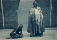 Le Mogho-Naba, Chef de tous les Mossis et un serviteur. Carte éditée par la Mission d’Ouagadougou vers 1920.  – « Hier, à Ouagadougou, j’ai été rendre visite au Moro-Naba, le roi de tout le pays Mossi. Il règne sur deux à trois millions de sujets et ses ancêtres furent en rapport avec les Portugais. M. Delafosse a même pu établir la chronologie des rois Mossi jusqu’à l’an mil. Le roi m’attend sur la porte de son palais, entouré de sa cour. Il est obèse ; une vraie outre noire, grasse ; l’air sensuel, féroce et malin. Barbiche, joues énormes. C’est bien le dernier roi nègre, celui de mes livres d’enfant. Il porte une énorme couronne d’or, une robe de velours violet soutachée d’or, et rafraîchit ses lourdes lèvres violacées d’une petite langue de carmin qui égaye de sa couleur tout le visage. Il me présente ses ministres, ses scribes, ses eunuques, ses pages, très beaux et vêtus de robes, coiffés en cimier, comme des femmes, comme elles guêtrés de jambières de cuivre de vingt centimètres de haut, parés de lourds anneaux de cuivre rose, d’une tonalité merveilleuse sur la peau nue. […] J’offre au Moro-Naba un bracelet d’émeraudes, acheté à Paris dans une de ces boutiques de faux à « tout pour cent francs… » (1). - Il doit s’agir du Mogho Naaba Koom II (1889 – 1942), l'arrière grand-père du Mogho Naaba actuel, qui régna du 27 février 1905 à sa mort, le 12 mars 1942. Il avait choisi pour devise « Que l'eau abonde pour tous ». Il repose, ainsi que sa femme, au centre de la place qui porte son nom devant la gare ferroviaire de Ouagadougou, sous la masse d'une statue métallique de 6 mètres de haut représentant une femme mossi stylisée offrant l'eau de bienvenue à un hôte invisible. Mogho Naaba Koom II s’était rallié à la France Libre dès le 22 juin 1940, alors que Paul Morand, dont on sent percer le dédain dans les lignes que lui a consacrées, sera nommé ambassadeur de France en Roumanie puis en Suisse par Vichy. Source : (1) Morand, Paul, A.O.F. de Paris à Tombouctou, Paris, Flammarion, 1928.