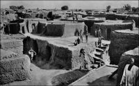 Quartier indigène de Bobo-Dioulasso. – Il s’agit d’un cliché de l’Agence économique des colonies qui situe la scène en Côte d’Ivoire. Cela signifie que la scène se situe dans l’intervalle entre 1932 et 1947, durant lequel la colonie de Haute-Volta a été intégrée aux colonies adjacentes. « Bobo-Dioulasso est une des ville les plus importante de la Côte d’Ivoire et forme la tête de ligne actuelle du chemin de fer qui remontera plus tard jusqu’à Ouagadougou… » (1). « Bobo est un gros centre cosmopolite : plus de 10 000 indigènes de toutes races s’y sont donné rendez-vous (Malinké, Bambara, Dioula, Mossi, Sia et Bobo). La gare domine la cité où émergent de-ci de-là quelques minarets. En sortant, nous voyons d’immenses entrepôts qui témoignent de l’activité commerciale de cette cité ; puis de larges avenues descendent en patte d’oie vers le centre de la ville, celle de droite nous conduit à la place de l’Etoile, à quelques mètre de laquelle nous apercevons les bâtiments de la mission des Pères Blancs » (2). « A Bobodioulasso, où nous couchons, nous trouvons la vraie capitale économique de la Haute-Volta. La ville est riche et se développe » (3). Sources : (1) Soubrier, J., Savanes et forêts, éditions J. Susse, Paris, 1944. (2) Paternot, Marcel, Lumière sur la Volta – Chez les Dagari, Lyon, Editions de la plus grande France, 1946. (3) Morand, Paul, A.O.F. de Paris à Tombouctou, Paris, Flammarion, 1928.