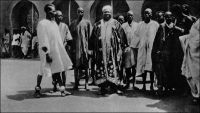 Le Moro-Naba, un des rois de la Haute-Volta, et sa cour. – « Hier, à Ouagadougou, j’ai été rendre visite au Moro-Naba, le roi de tout le pays Mossi. Il règne sur deux à trois millions de sujets et ses ancêtres furent en rapports avec les Portugais. M. Delafosse a même pu établir la chronologie des rois Mossi jusqu’à l’an mil. Le roi m’attend sur la porte de son palais, entouré de sa cour. Il est obèse ; une vraie outre noire, grasse ; l’air sensuel, féroce et malin. Barbiche, joues énormes. C’est bien le dernier roi nègre, celui de mes livres d’enfant. Il porte une énorme couronne d’or, une robe de velours violet soutachée d’or, et rafraîchit ses lourdes lèvres violacées d’une petite langue de carmin qui égaye de sa couleur tout le visage. Il me présente ses ministres, ses scribes, ses eunuques, ses pages, très beaux et vêtus de robes, coiffés en cimier, comme des femmes, comme elles guêtrés de jambières de cuivre de vingt centimètres de haut, parés de lourds anneaux de cuivre rose, d’une tonalité merveilleuse sur la peau nue. […] J’offre au Moro-Naba un bracelet d’émeraudes, acheté à Paris dans une de ces boutiques de faux à « tout pour cent francs… » (1). - Il doit s’agir du Mogho Naaba Koom II (1889 – 1942), l'arrière grand-père du Mogho Naaba actuel, qui régna du 27 février 1905 à sa mort, le 12 mars 1942. Il avait choisi pour devise « Que l'eau abonde pour tous ». Il repose, ainsi que sa femme, au centre de la place qui porte son nom devant la gare ferroviaire de Ouagadougou, sous la masse d'une statue métallique de 6 mètres de haut représentant une femme mossi stylisée offrant l'eau de bienvenue à un hôte invisible. Mogho Naaba Koom II s’était rallié à la France Libre dès le 22 juin 1940, alors que Paul Morand, dont on sent percer le dédain dans les lignes qui lui a consacrées, sera nommé ambassadeur de France en Roumanie puis en Suisse par Vichy. Source : (1) Morand, Paul, A.O.F. de Paris à Tombouctou, Paris, Flammarion, 1928.