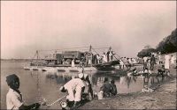 Fort-Lamy, l’arrivée du bac de Koussery. – « Lorsqu’on arrive à Fort-Lamy par le fleuve, on voit une ligne d’arbres vert sombre, dominés du drapeau français, ponctuée de flamboyants et cachant des maisons blanches. A cet endroit la rive du fleuve, aux basses eaux, est élevée comme une falaise. On croit à un paradis de verdure à l’abri des eaux. Avant ce promontoire de feuilles, la rive s’est abaissée en pente douce et sableuse ; les pirogues chargées de poissons, de bois, de paille, d’hommes, de femmes, d’enfants, d’animaux, de calebasses ; ouvertes de frêles, lourdes et couvertes de shimbecks de paille, grées de filets immenses et des apparaux de la pêche, ou nues ; de baleinières de métal, poussées par des perches ; un bac transportant un camion, c’est le port. » Source : Lapie, Pierre Olivier, Mes tournées au Tchad, Londres, John Murray, 1941.