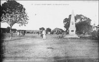 Fort-Lamy, la place du marché. – « L’entrée dans la ville est beaucoup plus décevante. Les concessions européennes sont minuscules, les jardins rares et pauvres, les arbres sur le bord du fleuve, les maisons ont un aspect sale et ruiné en raison de leur bâtisse de brique mauvaise que le soleil délite et de leur chaux locale, que la pluie délave. La place centrale est une sorte de désert où les enfants jouent au ballon, dans la poussière, les Européens au tennis ; les bicyclettes et les autos vont en tous sens et le marché indigène déploie ses paillotes, son odeur, ses incendies. […] Cette cité n’avait point d’ancêtre indigène. Fort-Lamy a été créé de toutes pièces après la victoire de Gentil sur Rabah. Les monuments de Gentil et Lamy, petits obélisques blancs entourés d’agaves ornent, si l’on peut dire, l’un la place, l’autre l’avenue du fleuve ». Source : Lapie, Pierre Olivier, Mes tournées au Tchad, Londres, John Murray, 1941.