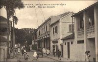 Dakar, rue des Essarts. – L’origine du nom de cette artère, l’une des plus anciennes de la capitale sénégalaise, fait débat. On s’accorde sur son intangibilité, elle s’est toujours appelée ainsi, de même qu’elle conserve de nos jours son tracé initial. Mais pour certains les « Essarts » font référence au défrichage nécessaire à son édification (c’est le nom donné à un espace gagné sur la forêt pour installer des activités humaines).  Tandis que pour d’autres, il s’agit d’un hommage à l’enseigne de vaisseau René Des Essart, tué par les troupes d’El Hadj Omar le 17 juillet 1857, lors du siège du Fort de Médine, à la veille de sa libération par le général Faidherbe. La rue fut tracée ex nihilo en 1862 - cinq ans seulement après l’installation française sur la presqu’île du Cap Vert - par le capitaine du génie Pinet-Laprade. Ce dernier organise le premier plan d’urbanisme de la ville, en s’inspirant du modèle d’une petite bourgade de province : il jette ainsi un boulevard portant son nom, quelques rues perpendiculaires et parallèles, dont la rue des Essarts, et une place centrale accueillant les activités de commerce, la place Kermel et le marché du même nom. Haut-lieu du quartier historique, la rue des Essarts accueillera, au fil du temps, le siège, la succursale locale ou la représentation pour l’AOF – dont Dakar était la capitale depuis 1902 - de nombreux opérateurs économiques et commerciaux, comme le Crédit Lyonnais, les magasins de confection « Ghislaine » et « Jaoudi », la CITEC – fournisseur officiel de « La toile d’avion » -, la Société anonyme de ravitaillement maritime… - Cette carte postale fait partie de l’importante collection due au photographe et éditeur dakarois Edmond Fortier (1862-1928), qui tenait d’ailleurs boutique rue Dagorne, une des voies transversales coupant la rue des Essarts au niveau du marché Kermel.