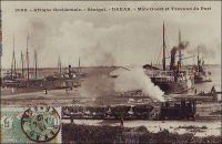 Dakar, Môle ouest et travaux du port – La ville de Dakar, devenue en 1902 la capitale de l’AOF, reçoit dès lors de forts investissements publics, pour développer les infrastructures tant civiles que militaires. Progressivement, le port qui est desservi par le chemin de fer à partir de1883, s’impose devant les autres installations du pays, et notamment Rufisque et Saint Louis. La machine à la manœuvre est la locomotive n°2 de type 030 T, fabriquée par les ateliers des Batignolles à Paris en 1883. – Cette photo fait partie du travail du célèbre photographe et éditeur dakarois Edmond Fortier (1862-1928) à qui l’on doit une somme importante de clichés sur l’Afrique de l’Ouest en général et sur le Sénégal en particulier ; il a publié en tout 3300 clichés originaux. M. Fortier tenait boutique et vivait avec ses deux filles blondes et son boy Seydou Traoré, à l’angle de la rue Dagorne et du boulevard Pinet-Laprade.