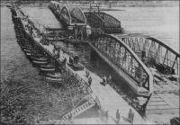Saint-Louis, pendant la construction du Pont Faidherbe. – Pendant la construit du pont métallique Faidherbe, inauguré en 1897, le pont de bateaux inauguré en 1865, et qui s’appelait déjà pont Faidherbe, reste en service. La traversée du grand bras du fleuve Sénégal, entre le quartier continental de Sor et l’île de Saint-Louis, était assurée depuis 1865 par un bac. Le nouveau pont Faidherbe, possède, comme le pont de bateaux avant lui, un point de passage pour les navires. En l’occurrence, la deuxième travée, à partir de la rive saint-louisiènne est mobile autour d’un pivot situé sur une pile. Le pont fait 511 mètres de longs, pour 10,50 mètres de large, avec une chaussée de 7 mètres et deux trottoirs piétons de 1,60 mètres. Ce projet avait été choisi pour son esthétique ; elle ne fut pas altérée par la restauration intervenue en 1931.