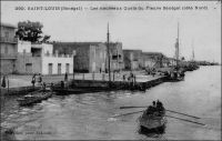 Saint-Louis, les nouveaux quais du fleuve Sénégal (côtés nord). - « La ville de Saint-Louis est bâtie sur une langue de sable, qu’on peut à la rigueur appeler une île, située presque au point de départ de deux bras du fleuve Sénégal, à environ douze mille de son embouchure. Cet îlot atteint à peine trois kilomètres de long, sur une largeur qui n’excède pas deux cents mètres ; sur toute l’étendue de la ville règnent des quais ou maçonneries bâtis sur pilotis. […] Saint-Louis communique par deux magnifiques ponts à la terre ferme, le pont fixe de Guet N’Dar sur le petit bras, et le pont de bateau mobile sur le grand bras, appelé pont de Sor. Des batteries bien installées défendent les approches de la ville de tous côtés, et une plantée à Guet N’Dar s’oppose à toute tentative de débarquement ». Source : Jacolliot, Louis, Voyage aux rives du Niger, Paris, C. Marpon et E. Flammarion, 1879. – Pierre Tacher (1875-1938), à qui l’on doit cette prise de vue, est un des pionniers de la photographie professionnelle au Sénégal. Son abondant travail, centré sur la région de Saint-Louis où il exerçait, est diffusé en cartes postales. Il était notamment connu pour son automobile, un tacot poussif, qui lui avait valu le surnom de « Borom Teuf-Teuf ». Il repose, avec son fils que l’on peut voir enfant sur une autre photo du site, au cimetière de Sor.