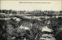Saint-Louis, panorama marché et pont. – Il s’agit ici du pont de Guet N’Dar sur le petit bras du fleuve Sénégal. « La ville de Saint-Louis est bâtie sur une langue de sable, qu’on peut à la rigueur appeler une île, située presque au point de départ de deux bras du fleuve Sénégal, à environ douze mille de son embouchure. Cet îlot atteint à peine trois kilomètres de long, sur une largeur qui n’excède pas deux cents mètres ; sur toute l’étendue de la ville règnent des quais ou maçonneries bâtis sur pilotis. […] Saint-Louis communique par deux magnifiques ponts à la terre ferme, le pont fixe de Guet N’Dar sur le petit bras, et le pont de bateau mobile sur le grand bras, appelé pont de Sor. Des batteries bien installées défendent les approches de la ville de tous côtés, et une plantée à Guet N’Dar s’oppose à toute tentative de débarquement ». Source : Jacolliot, Louis, Voyage aux rives du Niger, Paris, C. Marpon et E. Flammarion, 1879.