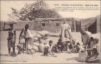 Boucle du Niger. Comptage des kawris dans un poste – Les kawris sont de petits coquillages servant de monnaie au Soudan (valeur moyenne 1 franc le mille). - Initialement utilisés comme monnaie par les populations des îles du continent indien (3000 ans avant JC), les kawris (ou cauris) sont introduits en Afrique noire entre le IXème et Xème siècle par le biais du commerce arabe transsaharien. Ils sont ensuite largement utilisés par les navigateurs portugais et espagnols dans les échanges avec le continent africain, et notamment pour la traite négrière. Pour la seule décennie qui va de 1850 à 1860, 35 000 tonnes de cauris auraient été importées en Afrique pour acquérir de l’huile de palme et des esclaves. Les types de coquillage et leur parité marchande varient selon les régions. Les cauris (Cypraea annulus, Cypraea moneta, Cypraea caurica, Cypraea onyx, Cypraea argus et Cypraea eglantina) ont cours. Les marginelles (Marginella persicula et Marginella cingulata) sont répandues au Soudan. Les olives (Olivella nana) sont utilisées au Congo. Dans les colonies françaises, la parité fluctuera, s’établissant un temps à 1000 cauris pour 1 francs, et jusqu’en 1907 les impôts peuvent être payés en coquillages au Soudan et au Dahomey, à raison de 20 000 cauris pour 7 francs. Par la suite les lois devaient interdire l’emploi des coquillages comme monnaie. Ils restèrent néanmoins utilisés dans des transactionslocales informelles, et au Niger à la fin des années 1940, on estimait à 70 000 cauris la dot due à la famille d’une épouse. Cette carte postale fait partie du travail d’Edmond Fortier (1862-1928), photographe et éditeur établis à Dakar. Elle doit dater de fin 1905, époque à laquelle il fit un voyage au Soudan.