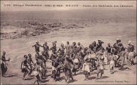 Macina, danse des habitants des cavernes. – Il s’agit vraisemblablement de Macina en tant que région, correspondant au royaume peul du XIX ème siècle, qui s’étendait de Tombouctou à Mopti et du Mossi à la Mauritanie. Quant aux habitants des cavernes, il doit s’agir des Habé, ou Habbé, désignation ancienne des Dogon de la région de Bandiagara. « Les Habés ont de curieuses danses. Les femmes n’y sont pas admises, de qui révèle leur caractère sacré. Les danseurs, au nombre de trente, forment un cercle. Ils sont nus, avec des rondelles de crins de cheval, d’une splendide couleur vieux-rose, autour de la taille, des poignets, des coudes et des chevilles ; ils sont coiffés d’une cagoule surmontée d’un cimier en coquillages blancs, cousus, dessinant des yeux ronds et vides. Leur danse est conduite par un meneur coiffé d’un masque d’oiseau, avec un long bec dentelé en scie, blanc et rouge ; l’homme emplumé ne cesse de déployer ses ailes, de siffler, simulacre d’un vrai oiseau qui gâte ici les récolte ; aussi ses danses sont-elles faites à une bonne distance des champs de mil » (1). Les traditions des Habbés, plus connus aujourd’hui sous le nom de Dogons, fascinent très tôt les voyageurs européens comme le photographe dakarois Edmond Fortier qui a immortalisé cette scène vers 1905. A leurs coutumes originales vient s’ajouter un peu plus tard la sulfureuse réputation de sauvagerie que leur vaut la révolte du village de Tabi en 1920, dernier épisode de résistance à la colonisation française, dans une région « pacifiée » lors des toutes dernières années du XIXème siècle. Haut perché sur la falaise de Bandiagara, le village constitue un bastion inexpugnable. Après sa prise, les coutumes troglodytes des Habbés sont interdites par le colonisateur. « La région est peuplée de Peulhs et surtout de Habés, population fétichiste assez arriérée… […] Elle vit en troglodyte, dans les montagnes de pierre feuilletée. […] On a interdit à ces Habés d’habiter leurs trous, où ils s’enfermaient après avoir retiré les échelle qui leur servaient à y monter. Maintenant ils ont construit de petits villages crénelés, dans des cirques de rochers rouges, au pied des falaises. L’administrateur du cercle me dit qu’il existe encore ici du cannibalisme rituel, et les Français rencontrés à Bandiagara me le confirment » (2). Sources (1) et (2) : Morand, Paul, A.O.F. de Paris à Tombouctou, Paris, Flammarion, 1928.