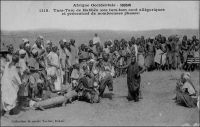 Tam-Tam de Habbés (ces tam-tam sont allégoriques et présentent de nombreuses phases). – « Les Habés ont de curieuses danses. Les femmes n’y sont pas admises, de qui révèle leur caractère sacré. Les danseurs, au nombre de trente, forment un cercle. Ils sont nus, avec des rondelles de crins de cheval, d’une splendide couleur vieux-rose, autour de la taille, des poignets, des coudes et des chevilles ; ils sont coiffés d’une cagoule surmontée d’un cimier en coquillages blancs, cousus, dessinant des yeux ronds et vides. Leur danse est conduite par un meneur coiffé d’un masque d’oiseau, avec un long bec dentelé en scie, blanc et rouge ; l’homme emplumé ne cesse de déployer ses ailes, de siffler, simulacre d’un vrai oiseau qui gâte ici les récolte ; aussi ses danses sont-elles faites à une bonne distance des champs de mil » (1). Les traditions des Habbés, plus connus aujourd’hui sous le nom de Dogons, fascinent très tôt les voyageurs européens comme le photographe dakarois Edmond Fortier qui a immortalisé cette scène vers 1905. A leurs coutumes originales vient s’ajouter un peu plus tard la sulfureuse réputation de sauvagerie que leur vaut la révolte du village de Tabi en 1920, dernier épisode de résistance à la colonisation française, dans une région « pacifiée » lors des toutes dernières années du XIXème siècle. Haut perché sur la falaise de Bandiagara, le village constitue un bastion inexpugnable. Après sa prise, les coutumes troglodytes des Habbés sont interdites par le colonisateur.  « La région est peuplée de Peulhs et surtout de Habés, population fétichiste assez arriérée… […] Elle vit en troglodyte, dans les montagnes de pierre feuilletée. […] On a interdit à ces Habés d’habiter leurs trous, où ils s’enfermaient après avoir retiré les échelle qui leur servaient à y monter. Maintenant ils ont construit de petits villages crénelés, dans des cirques de rochers rouges, au pied des falaises. L’administrateur du cercle me dit qu’il existe encore ici du cannibalisme rituel, et les Français rencontrés à Bandiagara me le confirment » (2). Sources (1) et (2) : Morand, Paul, A.O.F. de Paris à Tombouctou, Paris, Flammarion, 1928.