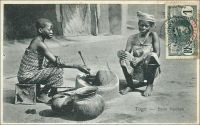 Togo - Beim Kochen (pendant la cuisson). – Cette carte postale, représentant une scène de cuisine dans le sud du Togo au tout début du XXe siècle, est intéressante à plusieurs titres. Elle témoigne en effet de ruptures et de continuités historiques et sociales. Ainsi, elle est issue d’une édition allemande, comme en témoigne la légende, et a donc été prise et publiée avant la première guerre mondiale. Pourtant, elle a été affranchie par un timbre français, donc expédiée après la prise de cette colonie allemande par les forces britannique et française en 1914, événement inaugural de la perte par l’Allemagne de tout son empire colonial. La scène elle-même raconte une vie encore organisée autour de productions locales. Les récipients de terre et de chaudronnerie traditionnelle, les ustensiles de bois et les bijoux sont de fabrications artisanales. Les textiles et la caisse servant de siège à la jeune cuisinière sont déjà des objets importés d’Europe. Aujourd’hui, ils seraient pour la plupart remplacés par des productions industrielles asiatiques. Même certains ingrédients seraient des produits d’importation, comme le bouillon en cube et le concentré de tomate, voire la viande surgelée. Seuls le lieu privilégié pour préparer le repas, la cour de la maison, et la cheville ouvrière de cette opération, une enfant vraisemblablement employée comme domestique, resteraient intangibles.