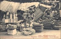 Porto-Novo, sur le marché. - « Le voyageur qui visite pour la première fois Porto-Novo est intéressé par le spectacle des rues. Celles-ci sont en effet très animées : on ne rencontre que femmes portant des caisses de genièvre, de muscat, manœuvres roulant des tonneaux de tafia, indigènes se rendant aux factoreries avec leurs pots d’huile de palme ou avec leur sac d’amande. Les marchandes à la criée ne sont pas inconnues. Chacun peut acheter, dans la rue, la nourriture habituelle de l’indigène : poisson fumé et bouteille d’acaça (farine de maïs bouillie). Dans les cuisines établies en plein vent, on vend au passant des friandises qui sortent toutes chaudes de l’huile de palme bouillante… » (1). Cette carte postale fait partie du travail du photographe et éditeur dakarois Edmond Fortier (1862-1928). Elle appartient à un reportage qu’il mena dans le sud du Dahomey en 1908, alors qu’il accompagnait, depuis l’escale de Dakar, le voyage officiel accompli sur la côte oust africaine par le Ministre des colonies Raphaël Milliès-Lacroix. Sources : (1)Hagen, A, « La colonie de Porto-Novo et le roi Toffa » dans, Revue d’ethnographie [ dir. Hamy, Ernest], Paris, éd. Ernest Leroux, 1887.