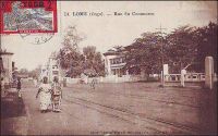 Lomé, rue du Commerce. - La rue du commerce est le centre d’affaire historique de la capitale togolaise. La ville s’est d’ailleurs constituée autour de sa vocation commerciale, à partir des années 1870 : des marchands Ewé, installés sur la côte pour mener la traite légitime (l’huile de palme essentiellement) avec les navires européens, sont venus se fixer en bordure de la frontière est de la Gold Coast, pour échapper aux lourdes taxes douanières britanniques. Aux pionniers se sont bientôt joints d’autres commerçants de la région, Mina venus de l’est et Haoussa descendus du nord. Au début des années 1880, des compagnies européennes, allemandes notamment, s’installent à Lomé, alors appelée Bey Beach (le nom du quartier de Bé en serait l’héritage). C’est sur l’initiative des commerçants allemands de Lomé – qui se sentaient légitimement menacés par les autorités coloniales anglaises, elles-mêmes exaspérées par cette concurrence à leurs portes - que l’explorateur Nachtigal s’arrête là, signe un traité et proclame le protectorat de l’empire allemand sur le Togo en 1884. Sur ce cliché, on peut distinguer, juste dans le dos des deux promeneuses, deux des hauts-lieux de la rue, la maison John Holt et tout à côté l’hôtel Gariglio, devenu ensuite Hôtel du Golfe.