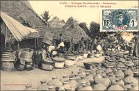 Porto-Novo, sur le marché. - « Le voyageur qui visite pour la première fois Porto-Novo est intéressé par le spectacle des rues. Celles-ci sont en effet très animées : on ne rencontre que femmes portant des caisses de genièvre, de muscat, manœuvres roulant des tonneaux de tafia, indigènes se rendant aux factoreries avec leurs pots d’huile de palme ou avec leur sac d’amande. Les marchandes à la criée ne sont pas inconnues. Chacun peut acheter, dans la rue, la nourriture habituelle de l’indigène : poisson fumé et bouteille d’acaça (farine de maïs bouillie). Dans les cuisines établies en plein vent, on vend au passant des friandises qui sortent toutes chaudes de l’huile de palme bouillante… » (1). Cette carte postale fait partie du travail du photographe et éditeur dakarois Edmond Fortier (1862-1928). Elle appartient à un reportage qu’il mena dans le sud du Dahomey en 1908, alors qu’il accompagnait, depuis l’escale de Dakar, le voyage officiel accompli sur la côte oust africaine par le Ministre des colonies Raphaël Milliès-Lacroix. Sources : (1)Hagen, A, « La colonie de Porto-Novo et le roi Toffa » dans, Revue d’ethnographie [ dir. Hamy, Ernest], Paris, éd. Ernest Leroux, 1887.