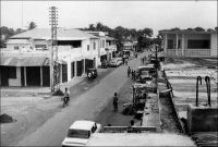Lomé, rue de l’église (dans les années 1960). – La capitale togolaise connait un fort développement à partir de la fin de la seconde Guerre mondiale. L’envolée du cours des produits tropicaux, les forts investissements publics et l’afflux de populations rurales, qui ont fuit les rigueurs de l’effort de guerre dans les campagnes, contribuent à l’explosion démographique et spatiale de la ville. Sa population bondit ainsi de 18 000 à 33 000 habitants entre 1938 et 1950, pour atteindre 85 000 habitants en 1958. Quittant la seule bande côtière, les quartiers s’étendent progressivement sur les vastes cocoteraies environnantes.  Vers le nord-ouest, sur les terres de la famille Olympio, un lotissement chic voit le jour et, plus loin, le quartier de Nyékonakpoé sort de terre. Vers le nord-est, Bé, son marché et sa gare – une zone jusqu’ici non prise en compte dans les dénombrements de la population loméenne – sont rejoints par les faubourgs. Vers le nord, la lagune est atteinte, puis franchie. Le plateau de Tokoin est progressivement investi, d’abord par les plus pauvres, puis par de grandes infrastructures publiques comme l’hôpital (l’actuel CHU sur le site de l’ancien terrain d’aviation) en 1954, le camp militaire, le collège Saint-Joseph entre 1948 et 1950… Parallèlement, la ville se modernise. Les biques d’argile, matière première justement prélevée sur le plateau de Tokoin et cuite avec les combustibles tirés des cocoteraies, cèdent le pas au parpaing de ciment dans la construction. L’électrification gagne du terrain, au-delà du quartier du wharf où elle avait débuté en 1926, et les rues sont goudronnées jusqu’au Boulevard Circulaire, notamment grâce aux crédits FIDES (Fonds d'investissement pour le développement économique et social d'outre-mer) crée en 1946.