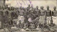 Adjiki, roi de Porto-Novo, ses ministres et sa cour. – Le roi Adjiki, monté sur le trône de Porto Novo en février 1908, connut une célébrité inédite dans la presse à sensation française. Peut être parce qu’il était le fils et le successeur du roi Toffa - l’allié de la France contre le roi Béhanzin d’Abomey à la fin du XIXème siècle – ou par nostalgie des choses de la royauté, son couronnement fut rapporté par Le Petit Journal Illustré, dans son édition du 5 avril 1908 : « Le prince Adjiki, fils de Toffa, coiffé du bicorne à plumes blanches, insigne de la souveraineté, assiste aux fêtes de son couronnement. Nous donnons plus loin, dans notre « variété », de curieux et pittoresques détails sur la façon dont on célébrait jadis, et dont on célèbre aujourd’hui, l’avènement d’un nouveau souverain dans ces contrées de la côte des Esclaves qui sont à présent colonies françaises.  C’est le récent décès de Toffa, roi de Porto-Novo, et le couronnement de son fils, le prince Adjiki, qui nous ont fourni l’occasion de donner à nos lecteurs, d’après des documents d’une absolue exactitude, cette intéressante gravure sur les fêtes qui se sont déroulées à Porto-Novo.  Adjiki a reçu le bicorne à plumes blanches, insigne du pouvoir, des mains de M. Marchal, lieutenant gouverneur de la colonie française du Dahomey, en même temps qu’un arrêté le nommait « chef supérieur des territoires français du Bénin ». De ce fait, Adjiki a pour attribution le concours à l’exécution de toutes les mesures prescrites par le lieutenant gouverneur ou par le président de Porto-Novo. Il intervient notamment lorsqu’il en est requis, auprès des chefs de région, de village ou de quartier pour les maintenir dans l’obéissance totale en toutes circonstances ou pour y maintenir les habitants du cercle de Porto-Novo. Il remplit les fonctions judiciaires qui peuvent ou pourront lui être dévolues par les actes régissant la matière.  Le prince Adjiki reçoit, en cette qualité de chef supérieur un traitement annuel de 25.000 francs, outre la part lui revenant des remises sur l’impôt personnel, allouées aux chefs  indigènes par les règlements en vigueurs. Et voilà comment il arrive parfois que la République fait des rois ».