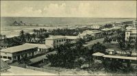 Lomé, Hamburger Strasse (vers 1905). – Connue à l’époque allemande sous le nom de Hamburgerstrasse, la rue du commerce est le centre d’affaire historique de la capitale togolaise. La ville s’est d’ailleurs constituée autour de sa vocation commerciale, à partir des années 1870 : des marchands Ewé, installés sur la côte pour mener la traite légitime (l’huile de palme essentiellement) avec les navires européens, sont venus se fixer en bordure de la frontière est de la Gold Coast, pour échapper aux lourdes taxes douanières britanniques. Aux pionniers se sont bientôt joints des commerçants Mina venus de l’est et Haoussa descendus du nord. Au début des années 1880, des compagnies européennes, allemandes notamment, s’installent à Lomé, alors appelée Bey Beach. C’est sur l’initiative des commerçants allemands de Lomé – qui se sentaient légitimement menacés par les autorités coloniales anglaises, elles-mêmes exaspérées par cette concurrence à leurs portes - que l’explorateur Nachtigal s’arrête là, signe un traité et proclame le protectorat de l’empire allemand sur le Togo en 1884. – Vraisemblablement prise depuis la galerie à l’étage de la mission catholique, cette photo montre la rue d’est vers l’ouest, c'est-à-dire schématiquement depuis la cathédrale vers le bâtiment de la douane allemande (démolie en 1983, pour laisser place à un centre commercial moderne comptant des salles de cinéma, un casino, une boite de nuit et un hôtel). On distingue au premier plan le toit de l’école catholique et à droite le terrain de boule, entouré de murs et où sera construit par la suite le bâtiment de l’UAC (United Africa Company). L’état du wharf, qui est achevé et ne compte que deux grues, situe la photo vers 1905 ; il sera par la suite rallongé de 50 m et doté de nombreux autres dispositifs de levage.