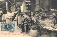 Porto-Novo, marchandes de colas et autres denrées indigènes. - « Le voyageur qui visite pour la première fois Porto-Novo est intéressé par le spectacle des rues. Celles-ci sont en effet très animées : on ne rencontre que femmes portant des caisses de genièvre, de muscat, manœuvres roulant des tonneaux de tafia, indigènes se rendant aux factoreries avec leurs pots d’huile de palme ou avec leur sac d’amande.Les marchandes à la criée ne sont pas inconnues. Chacun peut acheter, dans la rue, la nourriture habituelle de l’indigène : poisson fumé et bouteille d’acaça (farine de maïs bouillie). Dans les cuisines établies en plein vent, on vend au passant des friandises qui sortent toutes chaudes de l’huile de palme bouillante… » (1). Cette carte postale fait partie du travail du photographe et éditeur dakarois Edmond Fortier (1862-1928). Elle appartient à un reportage qu’il mena dans le sud du Dahomey en 1908, alors qu’il accompagnait, depuis l’escale de Dakar, le voyage officiel accompli sur la côte oust africaine par le Ministre des colonies Raphaël Milliès-Lacroix. Sources : (1)Hagen, A, « La colonie de Porto-Novo et le roi Toffa » dans, Revue d’ethnographie [ dir. Hamy, Ernest], Paris, éd. Ernest Leroux, 1887.