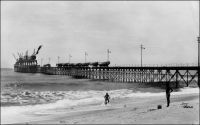 Lomé, le wharf. – Il s’agit du wharf français, bâti en 1928 pour se substituer au wharf allemand de 1904. Le nouvel édifice est construit perpendiculairement à la houle, et non plus à la plage, et possède une structure faite de légers croisillons métalliques offrant peu de résistance aux vagues. Ces précautions n’étaient pas superflues, sachant que le wharf allemand avait été partiellement emporté par la violence de la barre un jour de tempête en 1911. En 1954 le wharf est allongé de 50 m. et le nombre de ses grues porté de 6 à 10 ; la prise de vue est donc antérieure à cette adjonction. En 1962 le président Sylvanus Olympio procède à la pose de la première pierre du port en eau profonde de Lomé, appelé à remplacer le wharf. L’Imposante installation, qui entre en service en 1968, aura un impact environnemental conséquent, contribuant à l’ensablement de la côte occidentale et à l’érosion de la côte orientale.