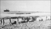 Lomé, frühere landung durch die Brecher.  (Débarquement d’autrefois à travers la barre), vers 1900. - Avant la construction du wharf par les Allemands, entre mars 1902 et janvier 1904, les marchandises, souvent conditionnées dans des tonneaux, étaient confiées aux vagues pour gagner la plage ; les pertes étaient importantes. La population de Lomé comptait, au dénombrement de 1900, quelques 111 Libériens, qui ne sont autres que les Krumen formant les équipages des baleinières et surfboats qui franchissaient la barre pour procéder à la manutention des marchandises auprès des navires au mouillage en rade foraine.