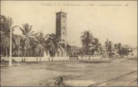 Lomé, le temple protestant. – Achevé en 1907, le temple subira en 1977 de profondes modifications architecturales et notamment l’amputation d’un étage du clocher. La photo a été prise après 1927, époque de l’électrification de la ville, on note en effet la présence d'un réseau électrique. – Les autorités coloniales allemandes s’emploient à maîtriser l’offre religieuse. Dans un premier temps elles fixent des zones d’installation aux différentes sociétés missionnaires, pour éviter qu’elles ne se disputent trop ostensiblement la conquête de nouveaux fidèles. Ainsi les catholiques n’eurent pas le droit de s’installer à Aného, alors la capitale. De plus, les autorités veillèrent à la germanisation des acteurs religieux ; le pasteur anglais d’Aného fut remplacé par un pasteur allemand. Il fut également négocié au plus haut niveau, pour que les missionnaires catholiques à venir s’installer au Togo soient allemands. A Lomé, les missions s’installent à partir de 1892, avec l’arrivée des pères et frères catholiques, qui bâtiront trois églises avant de construire la cathédrale entre 1901 et 1902. Les protestants, de la mission de Brème, installés dès 1853 à Keta, investissent Lomé en 1895 et y édifient le temple, pièce majeure de leur dispositif, entre 1906 et 1907.