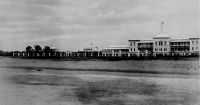 Lomé, Gouvernements-Gebäude. photo F. F. Olympio – Le quartier administratif (yovokomé) et le front de mer. L’imposant bâtiment est le logement administratif allemand (grosses Beantenwohnhaus), où étaient hébergés la plupart des agents supérieurs de la colonie jusqu’en 1908 ; construit entre 1897 et 1901, il est détruit dès 1913. Les fonctionnaires seront ensuite logés par deux dans des maisons de fonction, appelées « bungalow de fonctionnaires », toujours dans le même quartier. La vue est prise depuis le wharf allemand construit en 1903. Le dos de la carte postale n’est pas divisé, ce qui signifie qu’elle remonte à avant 1905 : la photo a donc vraisemblablement été prise en 1904. Fabriano Francisco Olympio, commerçant et photographe à Lomé, est l’un des fils de Francisco Olympio (le fondateur de la branche afro-brésilienne de la famille, né en 1833 à Bahia et arrivé sur la côte ouest africaine dans les années 1850) et le frère d’Octaviano Olympio, lui-même père de Sylvanus Olympio, le premier président du Togo indépendant. Ayant quitté le Togo au début du XXème siècle pour le Cameroun, Fabriano Francisco revint ensuite à Lomé ou il devait mourir en 1910.