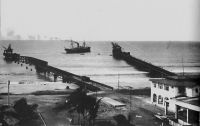 Lomé, la douane et les deux wharfs en 1928. – Le wharf allemand, à gauche sur l’image, fut construit entre 1902 et 1904, assurant l’essor économique de la ville qui était alors la seule dans toute la région à disposer d’une telle installation.  Mais une violente tempête, dans la nuit du 16 au 17 mai 1911, devait emporter la partie centrale de l’édifice, ne laissant que l’extrémité qui avait était ajoutée en 1908. Des travaux de restauration, entrepris en 1912,  aboutirent au contournement de la passerelle brisée, donnant au wharf réparé sa silhouette coudée si caractéristique. Le wharf français, à droite sur l’image, est construit entre 1925 et 1928. Profilé et perpendiculaire à la barre (et non plus à la côte comme le wharf allemand), il est plus résistant et offre une capacité de 700 tonnes par jour (le double du wharf allemand).